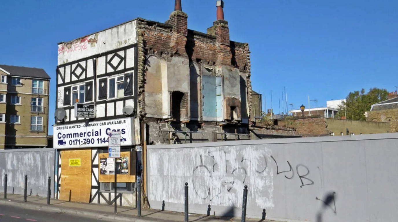 Existing derelict building on Poplar High Street in South Poplar with half-timbered facade and exposed brick chimneys surrounded by graffiti-covered fencing