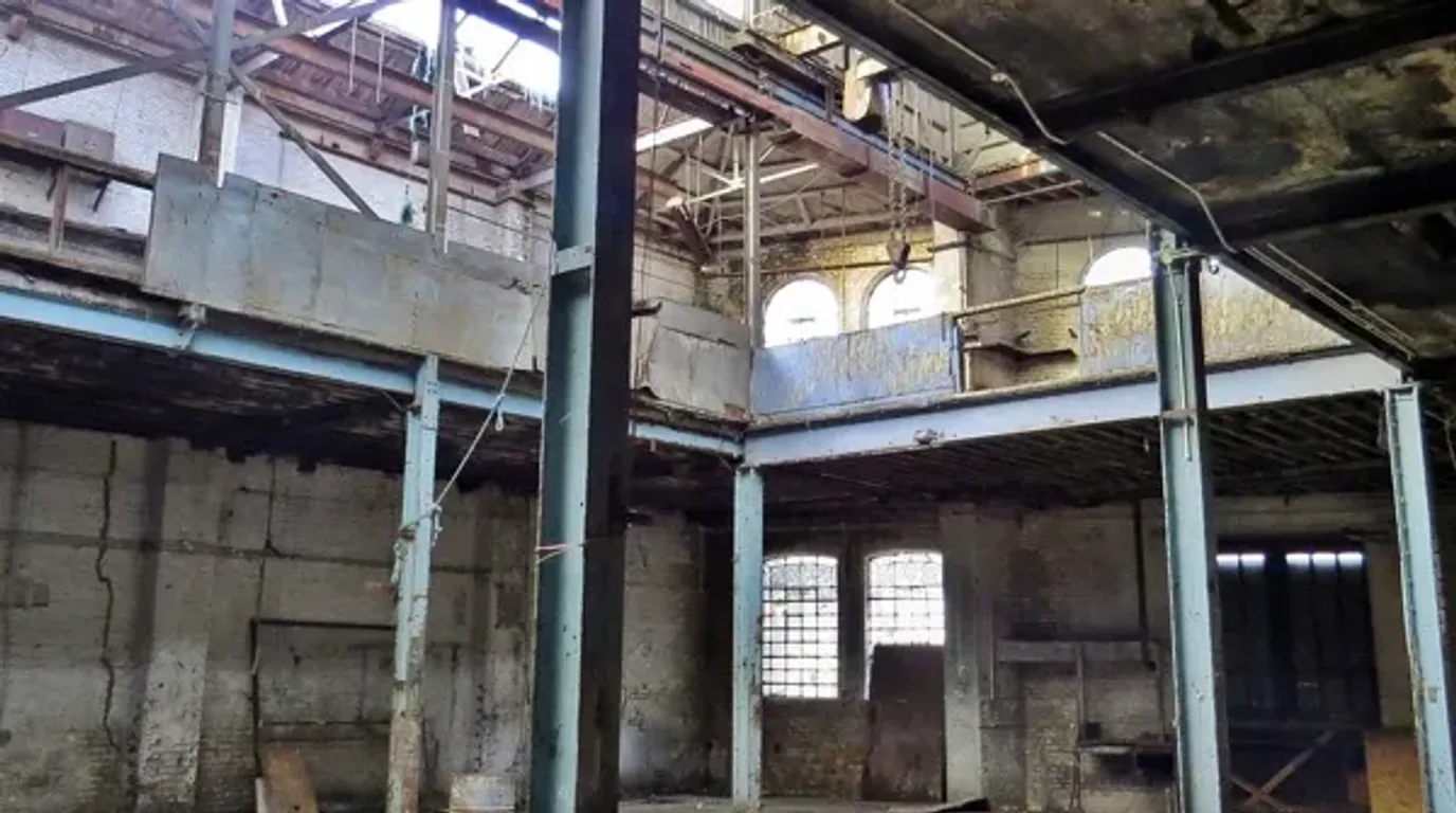 Existing derelict warehouse interior in South Poplar with exposed steel, vacant floor, mezzanine and damaged surfaces before adaptive reuse concept visualisation